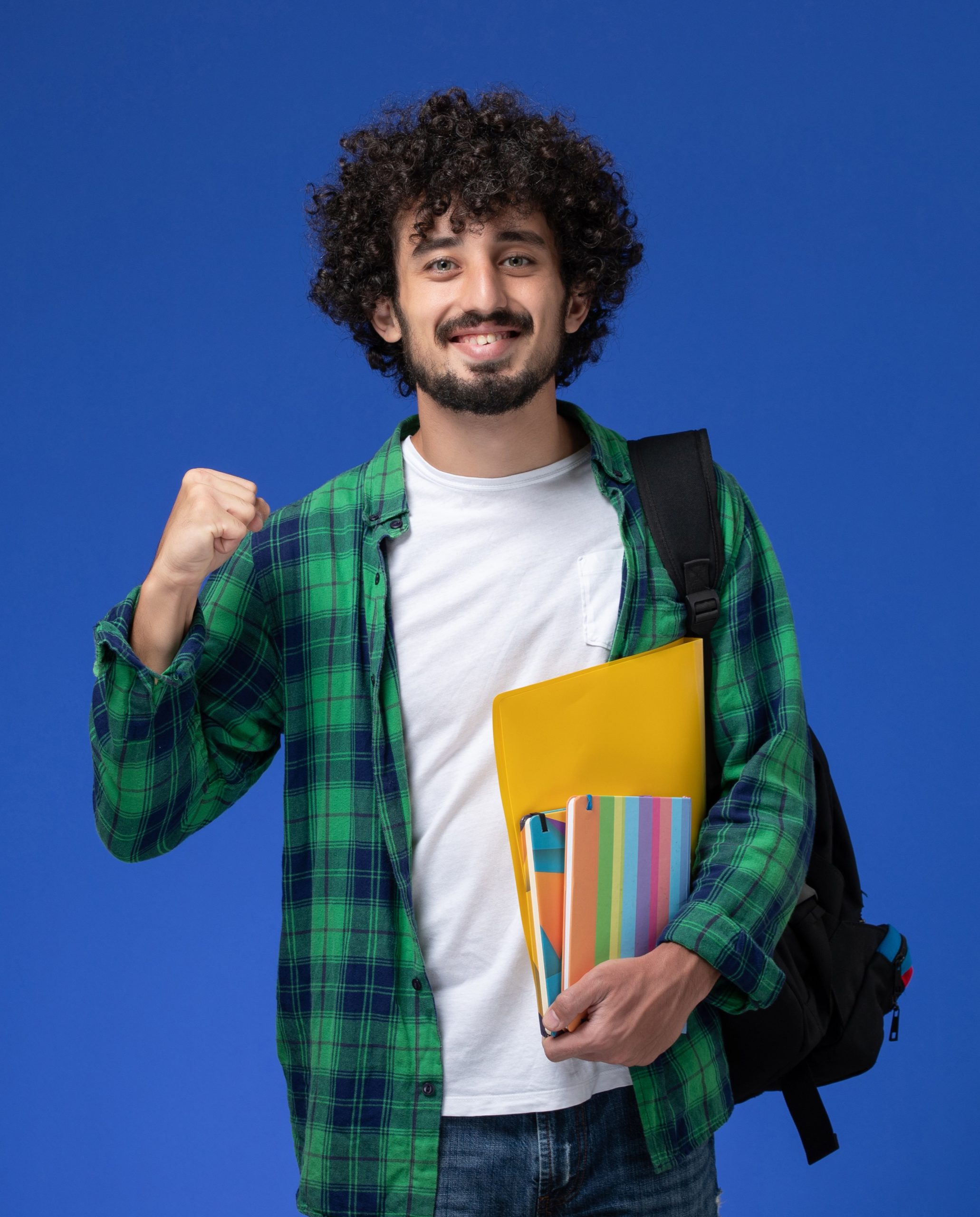 front view male student wearing black backpack holding copybooks files blue wall